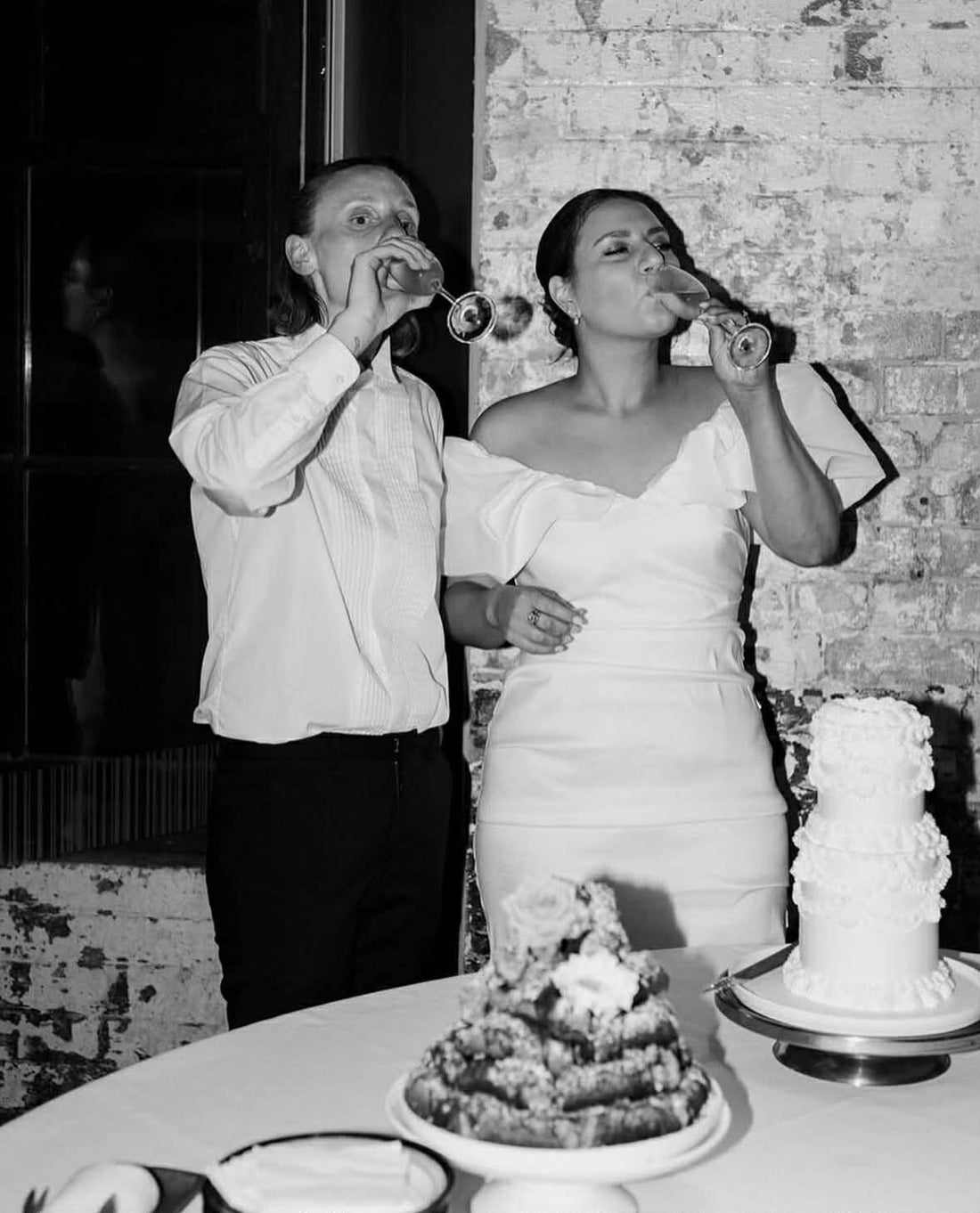 Man and woman in wedding attire standing next to a wedding cake, drinking from champagne glasses.