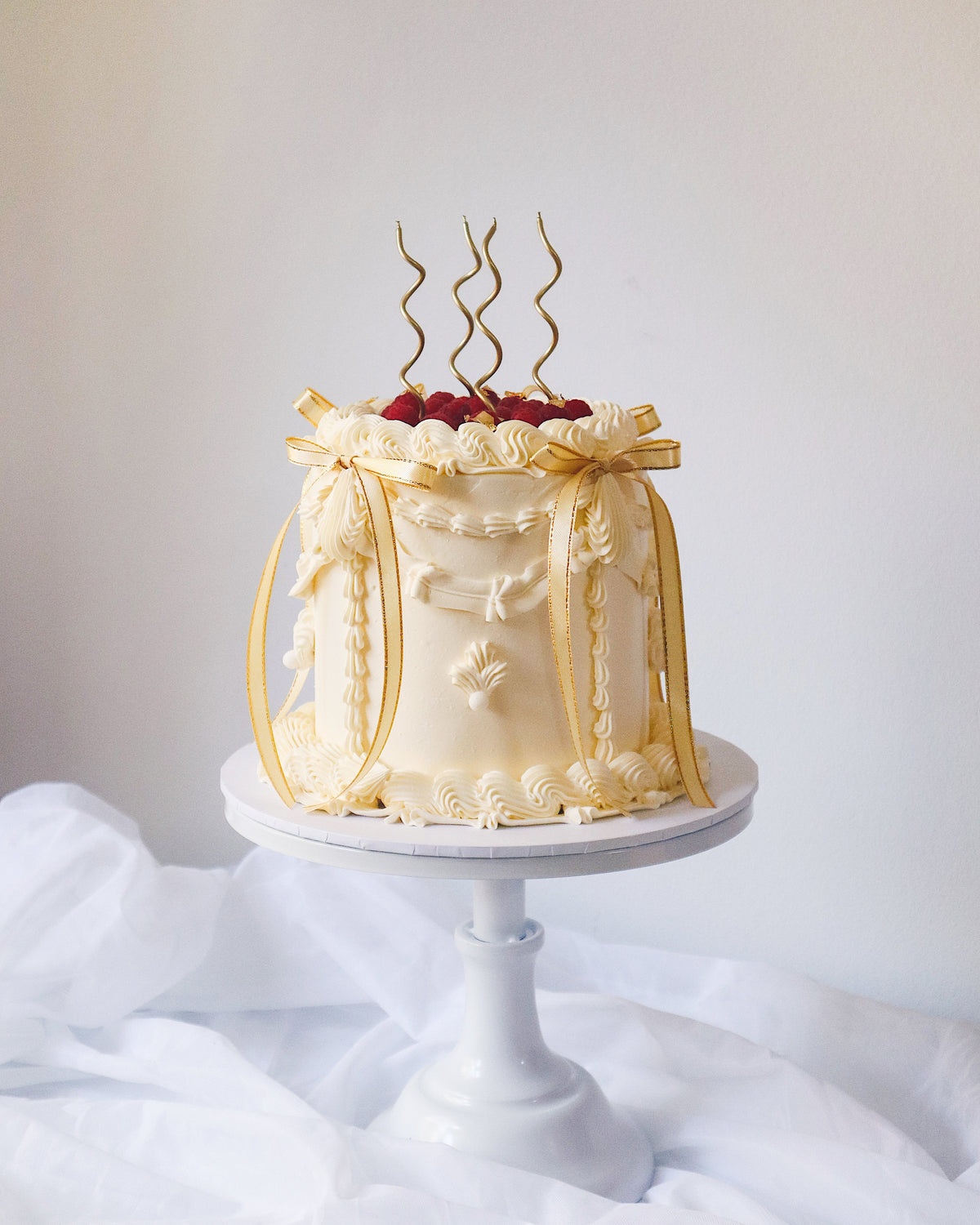 Decorative cake with white frosting and gold accents on a white stand against a light gray background.