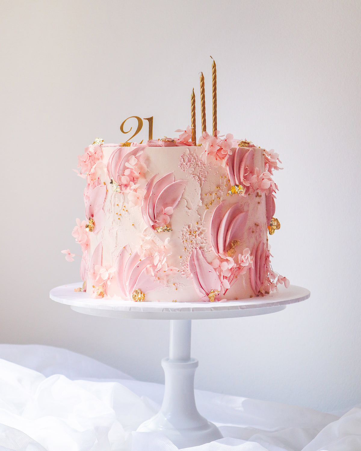 Pink cake with floral decorations and gold candles on a white stand against a light background