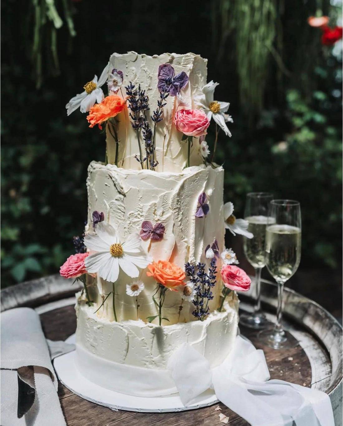 Three-tiered wedding cake with floral decorations on a wooden table outdoors.