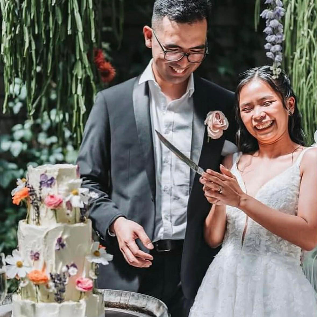 Couple cutting a wedding cake with a knife, surrounded by greenery.