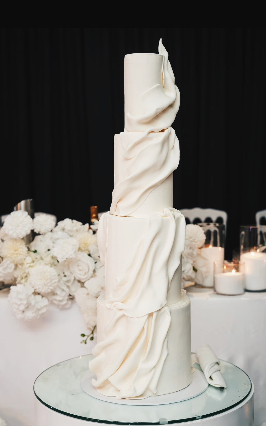 Elegant white wedding cake with ruffled details on a table with floral decorations.