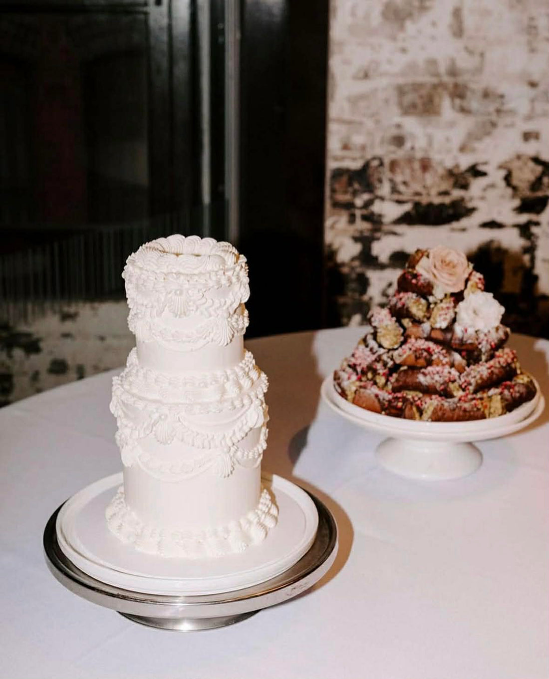 Three-tiered white wedding cake and a smaller dessert on a table with a brick wall background.